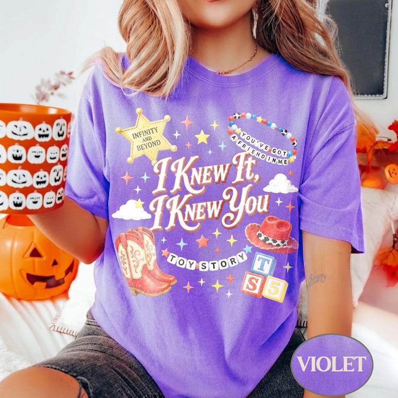 May include: A violet short-sleeve t-shirt featuring a graphic print with the text "I Knew It, I Knew You" and "Toy Story". The design includes a yellow sheriff star, a beaded bracelet, a red cowboy hat, cowboy boots, and alphabet blocks.