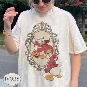以下が含まれることがあります： アイボリーの半袖Tシャツ。赤いマントとフードを身につけ、ジャック・オー・ランタンのバケツを持った漫画のキャラクターのダックが描かれています。グラフィックは、茶色の装飾的なヴィンテージ風の楕円形のフレームで囲まれています。