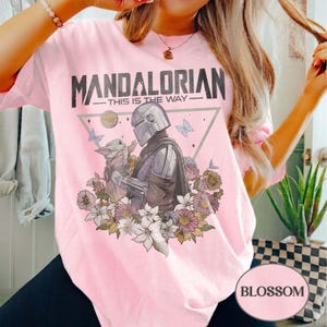 Op de afbeelding: Zachtroze T-shirt met een afbeelding van The Mandalorian en Grogu, omringd door bloemen en vlinders. De tekst "MANDALORIAN - THIS IS THE WAY" staat boven de afbeelding. Het shirt heeft een ronde hals en korte mouwen.
