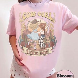 Puede incluir: Una camiseta rosa claro con una ilustraci&oacute;n gr&aacute;fica de dos figuras con sombreros de vaquero y botas en un paisaje des&eacute;rtico con cactus y rosas. El texto COW GIRL y LIKE ME est&aacute; impreso en una fuente decorativa sobre y debajo de las figuras.