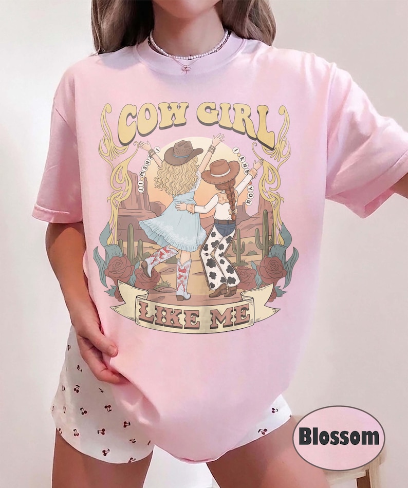 Puede incluir: Una camiseta rosa claro con una ilustraci&oacute;n gr&aacute;fica de dos figuras con sombreros de vaquero y botas en un paisaje des&eacute;rtico con cactus y rosas. El texto COW GIRL y LIKE ME est&aacute; impreso en una fuente decorativa sobre y debajo de las figuras.