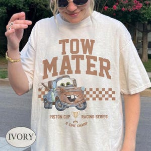 May include: An ivory t-shirt featuring the brown text TOW MATER in a collegiate font above a graphic of the character Tow Mater from the movie Cars. Below the character is a checkered pattern, the text PISTON CUP RACING SERIES, and 5 TIME CHAMP.