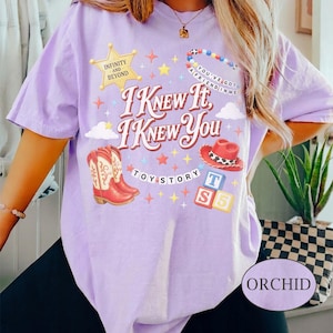 May include: A light purple t-shirt featuring a graphic print with the text I KNEW IT, I KNEW YOU, TOY STORY, INFINITY AND BEYOND, and YOU'VE GOT A FRIEND IN ME. The design includes a yellow star badge, red cowboy boots, a red hat, and alphabet blocks.