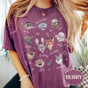 May include: A berry-colored short-sleeve t-shirt featuring a graphic print of various animated animal characters from Zootopia, including Judy Hopps, Nick Wilde, and Flash, with their names written in white script and small star accents throughout.