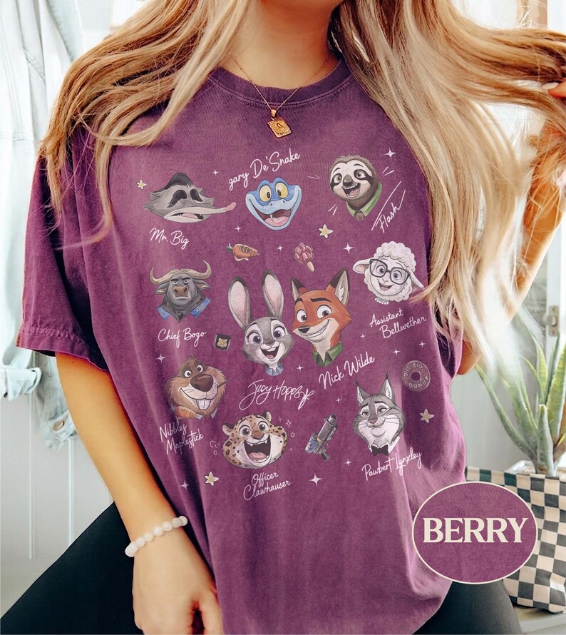 May include: A berry-colored short-sleeve t-shirt featuring a graphic print of various animated animal characters from Zootopia, including Judy Hopps, Nick Wilde, and Flash, with their names written in white script and small star accents throughout.