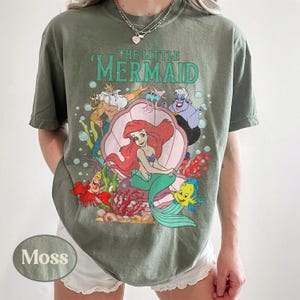 May include: Moss green t-shirt featuring a graphic from Disney's The Little Mermaid. The design includes Ariel, Flounder, Sebastian, King Triton, and Ursula. The words "THE LITTLE MERMAID" are printed above the characters. The shirt is a relaxed fit.