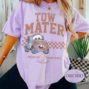 May include: A light purple t-shirt featuring a graphic of the character Tow Mater from the movie Cars. The design includes the text TOW MATER, a checkered pattern, and the words PISTON CUP RACING SERIES 5 TIME CHAMP with a trophy icon.