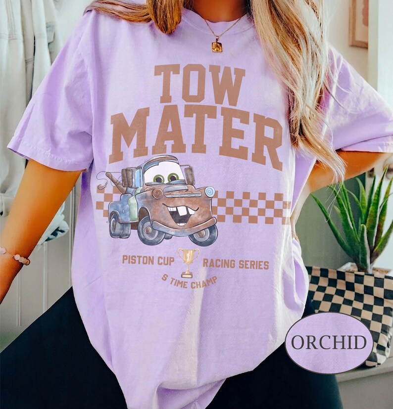 May include: A light purple t-shirt featuring a graphic of the character Tow Mater from the movie Cars. The design includes the text TOW MATER, a checkered pattern, and the words PISTON CUP RACING SERIES 5 TIME CHAMP with a trophy icon.
