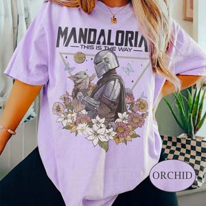 Op de afbeelding: Lichtpaars T-shirt met de tekst "MANDALORIA" en de zin "THIS IS THE WAY". Het ontwerp bevat een illustratie van The Mandalorian en Grogu, omringd door bloemen en vlinders. Het shirt heeft korte mouwen en een losse pasvorm.