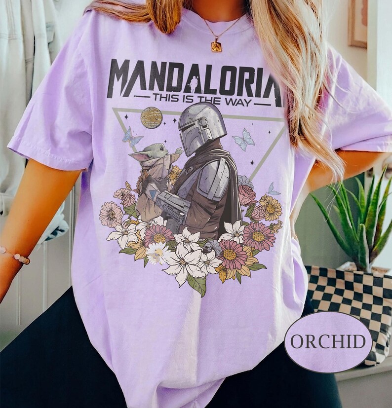 Op de afbeelding: Lichtpaars T-shirt met de tekst "MANDALORIA" en de zin "THIS IS THE WAY". Het ontwerp bevat een illustratie van The Mandalorian en Grogu, omringd door bloemen en vlinders. Het shirt heeft korte mouwen en een losse pasvorm.