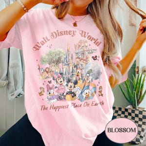 May include: Pink t-shirt featuring a colorful graphic of Disney characters and landmarks, with the text "Walt Disney World" and "The Happiest Place On Earth". The design includes Mickey Mouse, Minnie Mouse, Donald Duck, and other characters.