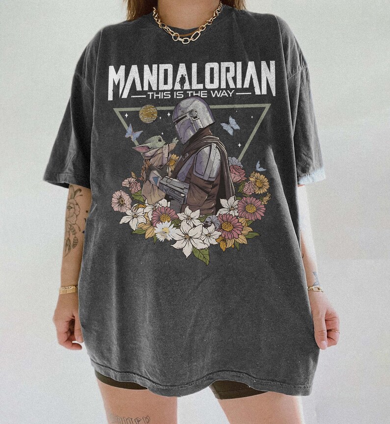 Op de afbeelding: Donkergrijze t-shirt met een afbeelding van The Mandalorian en Grogu, omringd door bloemen. De tekst "MANDALORIAN THIS IS THE WAY" staat bovenaan. Het t-shirt heeft korte mouwen en een losse pasvorm.