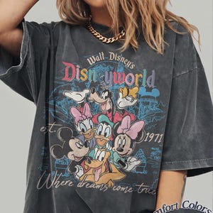 May include: Dark gray t-shirt featuring a vintage-style Walt Disney World graphic with Mickey Mouse, Minnie Mouse, Donald Duck, Daisy Duck, Goofy, and Pluto. The shirt has the text "Where dreams come true" and "est. 1971". The shirt is labeled "Comfort Colors Pepper".