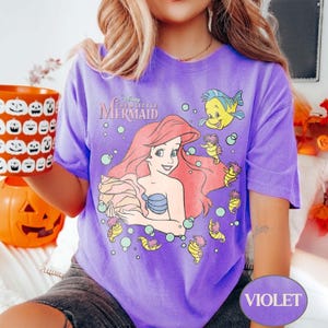May include: A violet t-shirt featuring a graphic of Ariel from Disney's The Little Mermaid. The design includes Ariel holding a seashell, with Flounder and seahorses. The shirt has the text "Disney The Little Mermaid".