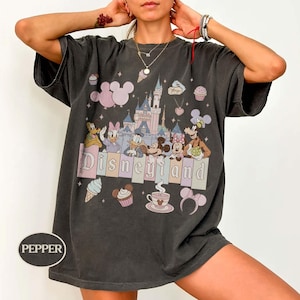 Może przedstawiać: Ciemnoszara koszulka z grafiką Disneyland. Projekt zawiera postacie z kresk&oacute;wek, zamek i napis "Disneyland" w pastelowych kolorach. Dodatkowe elementy to rożki lod&oacute;w, babeczki i sylwetki Myszki Miki.