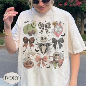 May include: An ivory short-sleeve t-shirt featuring a graphic print of characters from The Nightmare Before Christmas, including Jack Skellington, Sally, Oogie Boogie, and Zero, interspersed with various patterned bows and spider web motifs.