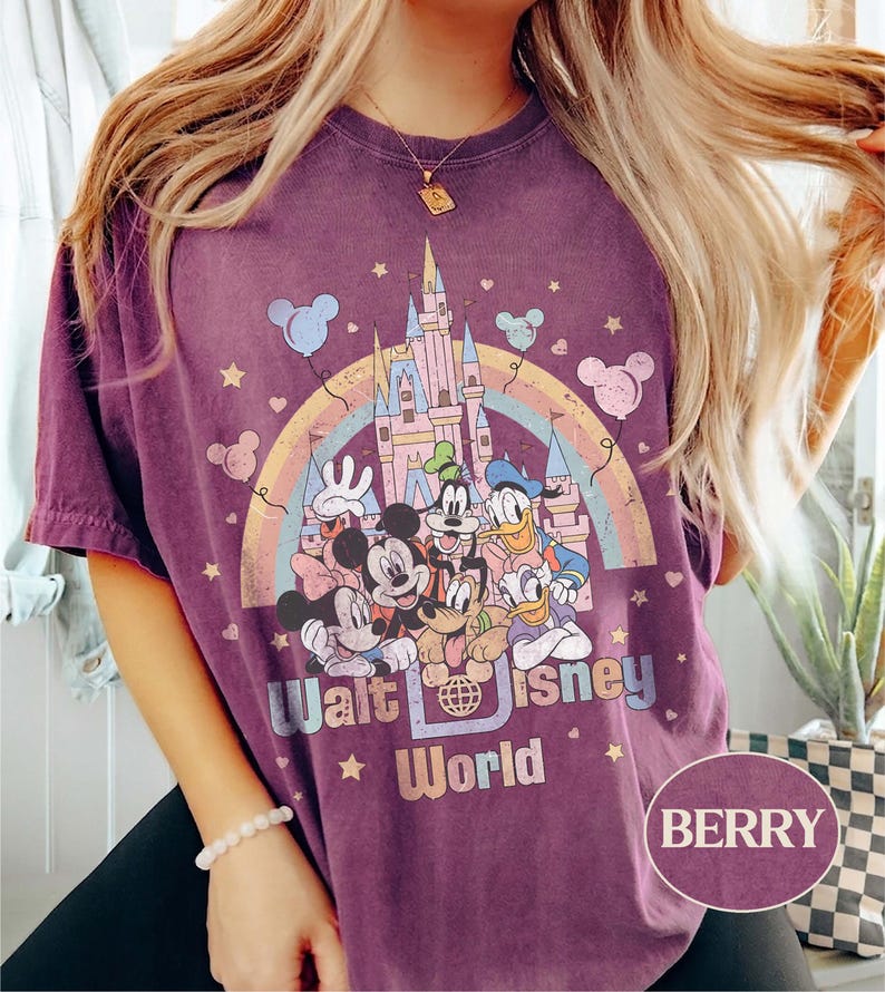May include: Berry-colored t-shirt featuring a colorful graphic of Disney characters, a castle, and a rainbow. The text "Walt Disney World" is printed below the characters. The shirt has short sleeves.