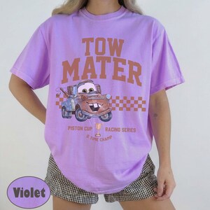 May include: A light purple short-sleeve t-shirt featuring a graphic of the character Tow Mater. The text reads TOW MATER, PISTON CUP RACING SERIES, and 5 TIME CHAMP. A brown checkered pattern sits behind the character, and a trophy icon is included.