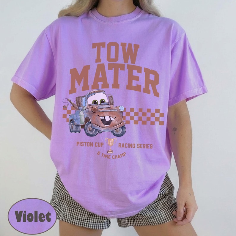 May include: A light purple short-sleeve t-shirt featuring a graphic of the character Tow Mater. The text reads TOW MATER, PISTON CUP RACING SERIES, and 5 TIME CHAMP. A brown checkered pattern sits behind the character, and a trophy icon is included.