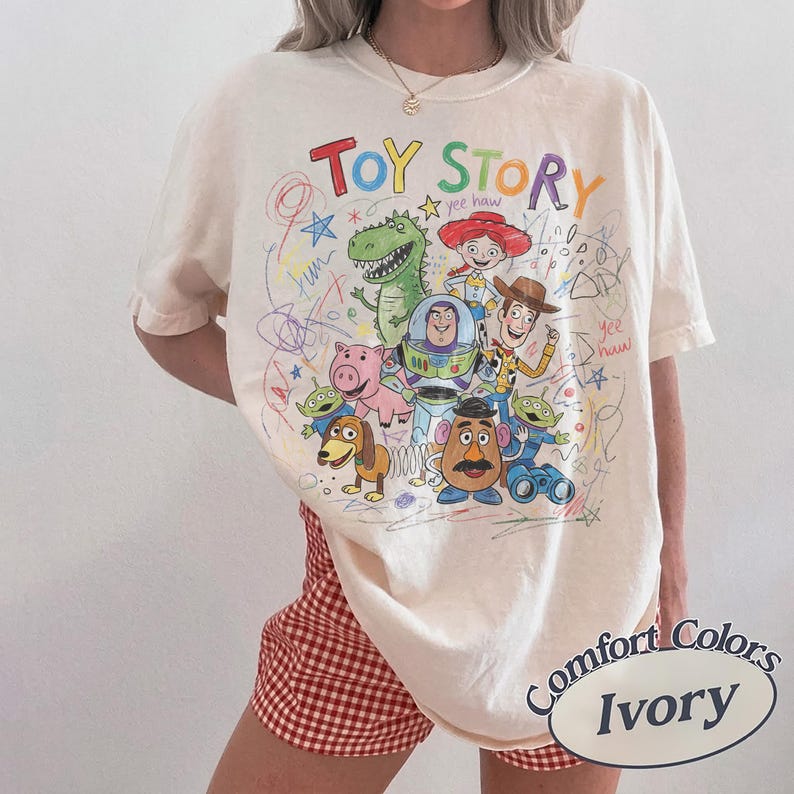 Pode incluir: Camiseta de cor marfim com um gr&aacute;fico colorido dos personagens de Toy Story, incluindo Woody, Buzz Lightyear e Rex. As palavras "TOY STORY" est&atilde;o escritas em um arco-&iacute;ris acima dos personagens. A camiseta &eacute; da marca Comfort Colors.