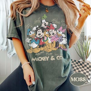 K&ouml;nnte beinhalten: Moosgr&uuml;nes T-Shirt mit einer Grafik von Micky Maus und ihren Freunden. Das Design umfasst Micky, Minnie, Donald, Daisy, Goofy und Pluto. Der Text "Mickey & Co. Est. 1928" ist darunter gedruckt.