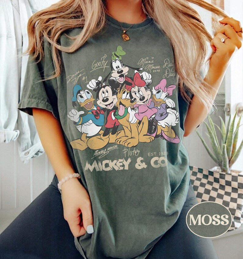 K&ouml;nnte beinhalten: Moosgr&uuml;nes T-Shirt mit einer Grafik von Micky Maus und ihren Freunden. Das Design umfasst Micky, Minnie, Donald, Daisy, Goofy und Pluto. Der Text "Mickey & Co. Est. 1928" ist darunter gedruckt.