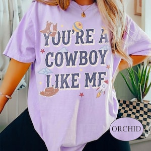 May include: A light purple short-sleeve t-shirt featuring the text "YOU'RE A COWBOY LIKE ME" in dark blue letters. The design includes illustrations of cowboy boots, a hat, a ball, a rocket, stars, and clouds scattered around the text.