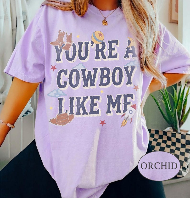 May include: A light purple short-sleeve t-shirt featuring the text "YOU'RE A COWBOY LIKE ME" in dark blue letters. The design includes illustrations of cowboy boots, a hat, a ball, a rocket, stars, and clouds scattered around the text.