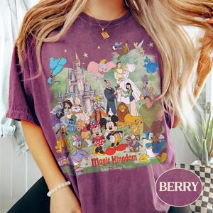 May include: Berry-colored t-shirt featuring a colorful graphic of Disney characters and the Magic Kingdom castle. The design includes Mickey Mouse, Donald Duck, and other beloved characters. The text "Magic Kingdom Walt Disney World" is printed below the characters.