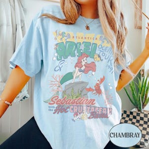 May include: A light blue t-shirt featuring a graphic print of Ariel, Flounder, and Sebastian from The Little Mermaid. The text reads Atlantica Ocean Tour 1991, Ariel, and Sebastian with the Hot Crustacean Band, surrounded by coral and bubbles.