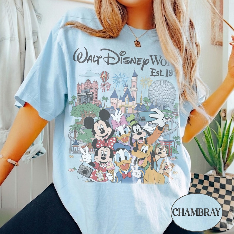 May include: A light blue t-shirt featuring a graphic print of Walt Disney World with the text "Walt Disney World Est. 1971". The illustration includes Mickey Mouse, Minnie Mouse, Donald Duck, Daisy Duck, Goofy, and Pluto in front of park landmarks.