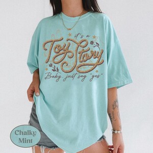 May include: A chalky mint green short-sleeve t-shirt featuring a brown rope-style graphic that reads "it's a Toy Story Baby, just say "yes"" in cursive script, accented with small stars and floral illustrations on the front center.