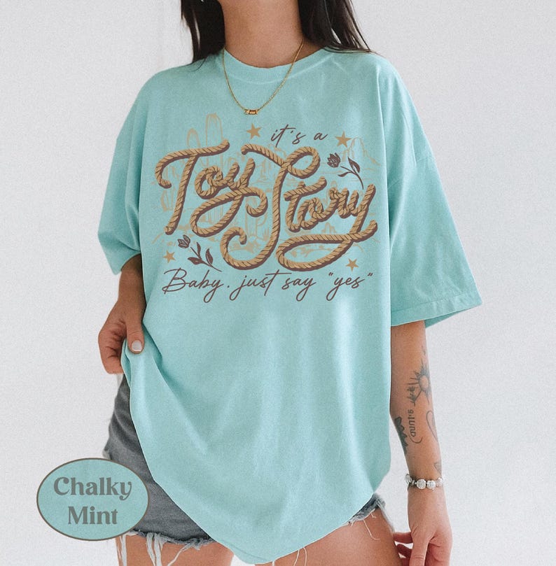 May include: A chalky mint green short-sleeve t-shirt featuring a brown rope-style graphic that reads "it's a Toy Story Baby, just say "yes"" in cursive script, accented with small stars and floral illustrations on the front center.