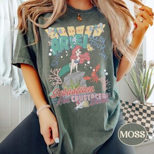 May include: A dark green short-sleeve t-shirt featuring a graphic print of Ariel, Flounder, and Sebastian from The Little Mermaid. The text reads Atlantica Ocean Tour 1991, Sebastian with the Hot Crustacean Band, and Ariel in a vintage style.