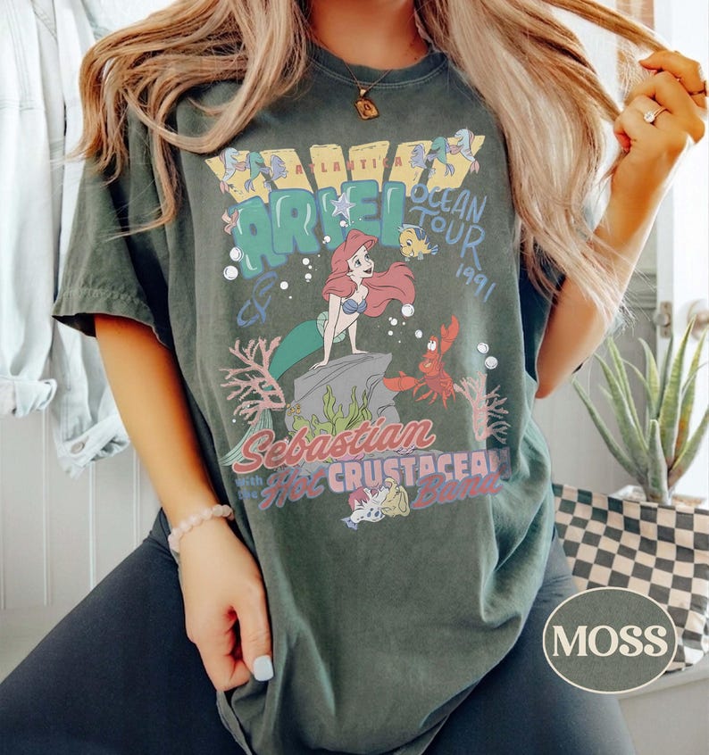 May include: A dark green short-sleeve t-shirt featuring a graphic print of Ariel, Flounder, and Sebastian from The Little Mermaid. The text reads Atlantica Ocean Tour 1991, Sebastian with the Hot Crustacean Band, and Ariel in a vintage style.