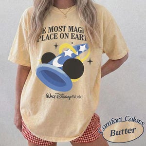 Puede incluir: Camiseta amarilla clara con un dise&ntilde;o de un sombrero de mago azul y blanco, orejas negras de Mickey Mouse y el texto "THE MOST MAGICAL PLACE ON EARTH" y "Walt Disney World". La camiseta es de color Mantequilla Comfort Colors.