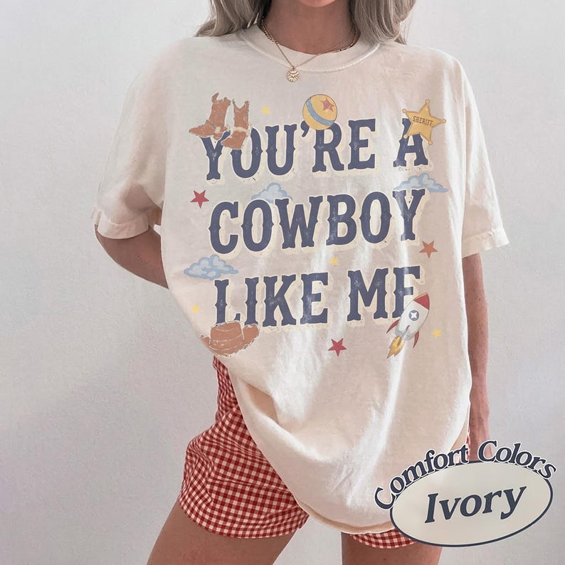 May include: An ivory short-sleeve t-shirt featuring the dark blue text "YOU'RE A COWBOY LIKE ME" surrounded by graphic illustrations of brown cowboy boots, a yellow ball, a sheriff star, blue clouds, red stars, and a rocket ship.