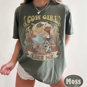 Puede incluir: Una camiseta de manga corta color verde musgo con un estampado gr&aacute;fico de dos figuras con sombreros de vaquero bailando en un paisaje des&eacute;rtico. El texto "COW GIRL" aparece arriba y "LIKE ME" abajo, enmarcado por ilustraciones de flores y cactus.