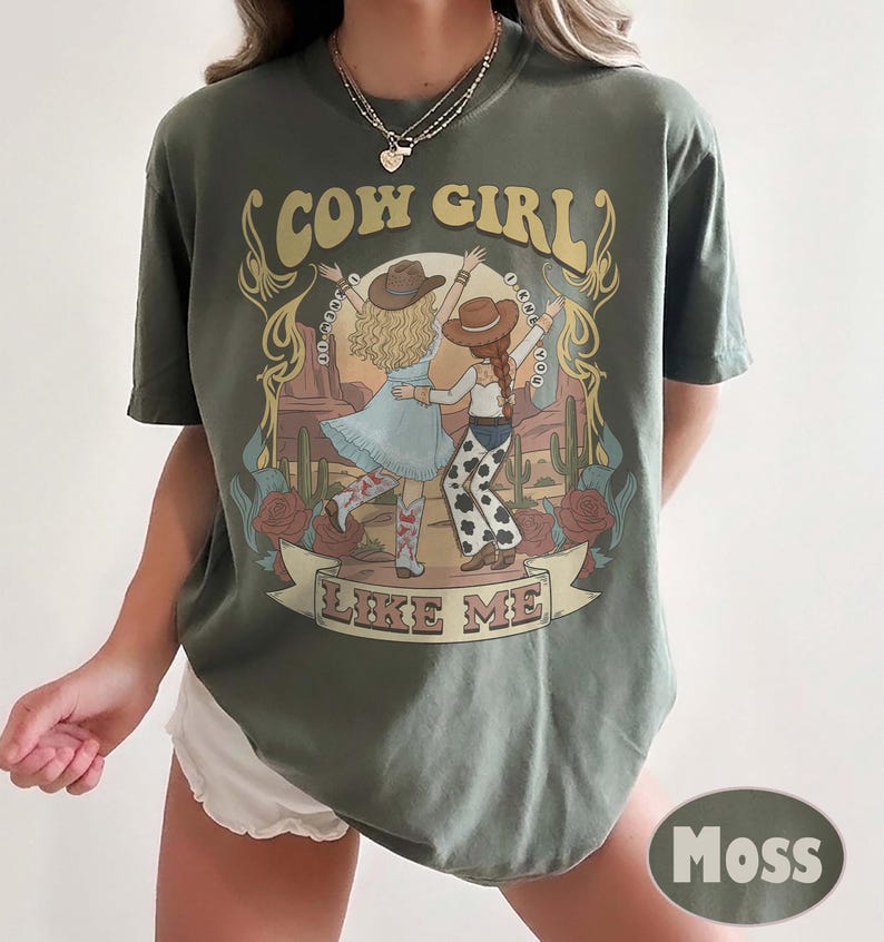 Puede incluir: Una camiseta de manga corta color verde musgo con un estampado gr&aacute;fico de dos figuras con sombreros de vaquero bailando en un paisaje des&eacute;rtico. El texto "COW GIRL" aparece arriba y "LIKE ME" abajo, enmarcado por ilustraciones de flores y cactus.