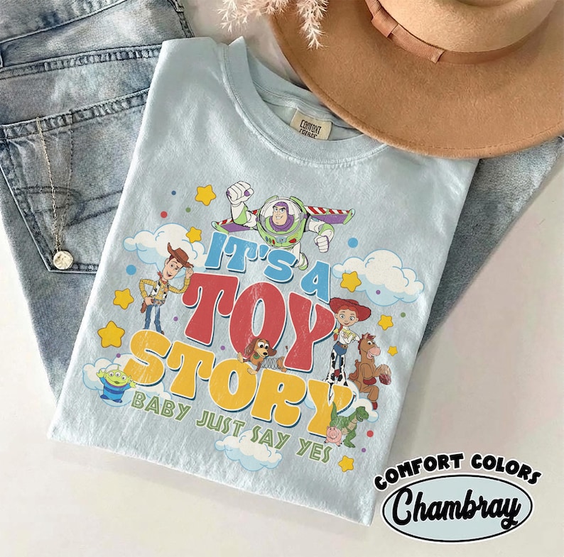 Peut inclure: Un t-shirt bleu clair avec une impression graphique des personnages de Toy Story, dont Woody, Buzz l'&Eacute;clair et Jessie. Le texte indique It's A Toy Story Baby Just Say Yes. Le t-shirt est pr&eacute;sent&eacute; avec un jean bleu, un chapeau beige et un collier.