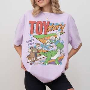 May include: A light purple t-shirt featuring a graphic of Toy Story characters, including Woody, Buzz Lightyear, and Rex the dinosaur. The words "TOY STORY" are in red and orange, with "SINCE 1995" below. The shirt has short sleeves.