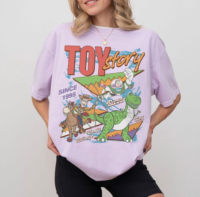 May include: A light purple t-shirt featuring a graphic of Toy Story characters, including Woody, Buzz Lightyear, and Rex the dinosaur. The words "TOY STORY" are in red and orange, with "SINCE 1995" below. The shirt has short sleeves.