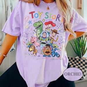 Pode incluir: Uma camiseta lil&aacute;s clara com um gr&aacute;fico colorido de personagens do filme de anima&ccedil;&atilde;o "Toy Story". O texto "Toy Story" est&aacute; no topo, com as palavras "Yee Haw" abaixo. A camiseta tem mangas curtas e um corte folgado.