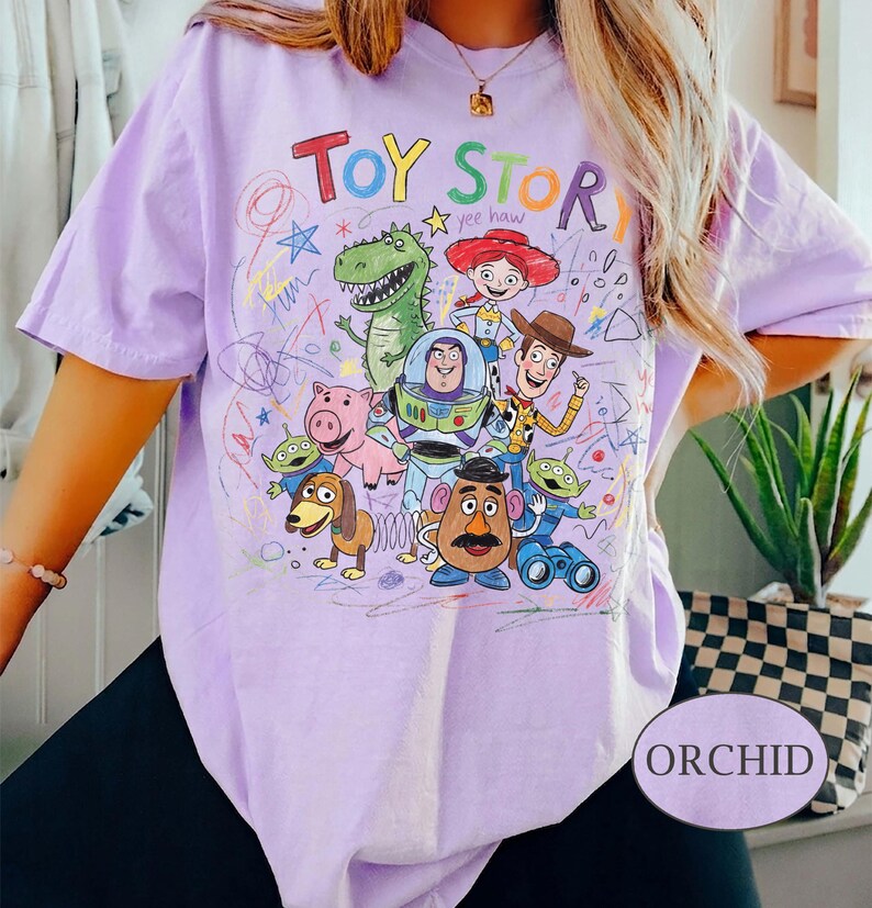 Pode incluir: Uma camiseta lil&aacute;s clara com um gr&aacute;fico colorido de personagens do filme de anima&ccedil;&atilde;o "Toy Story". O texto "Toy Story" est&aacute; no topo, com as palavras "Yee Haw" abaixo. A camiseta tem mangas curtas e um corte folgado.