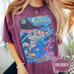 Puede incluir: Camiseta de color bayas con un gr&aacute;fico de Buzz Lightyear de la pel&iacute;cula Toy Story. El dise&ntilde;o incluye las palabras "Buzz Lightyear Laser Blast" y "Discoveryland" con im&aacute;genes de los personajes en una escena espacial.