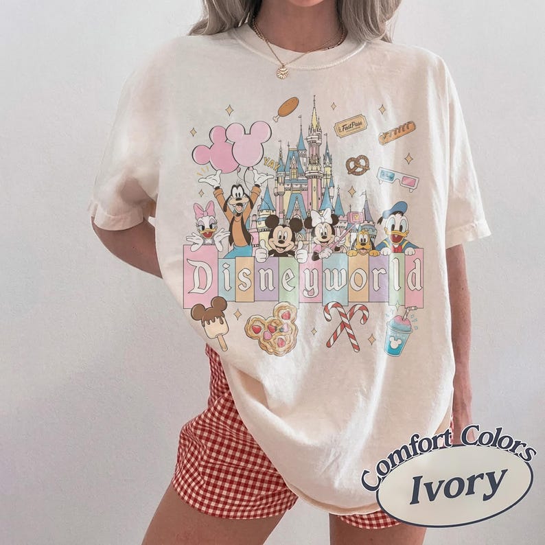 May include: Ivory-coloured t-shirt with a colourful Disney World graphic. The design features Mickey Mouse, Minnie Mouse, Donald Duck, and other characters in front of a castle. The words "Disneyworld" are displayed in pastel colours. The shirt is a Comfort Colors brand.