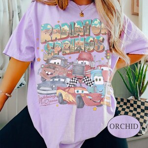 K&ouml;nnte beinhalten: Orchideenfarbenes T-Shirt mit einer Grafik von Charakteren aus dem Film Cars, darunter Lightning McQueen. Das Design enth&auml;lt den Text "Radiator Springs" und "Lightning McQueen". Das Shirt hat kurze &Auml;rmel und einen Rundhalsausschnitt.