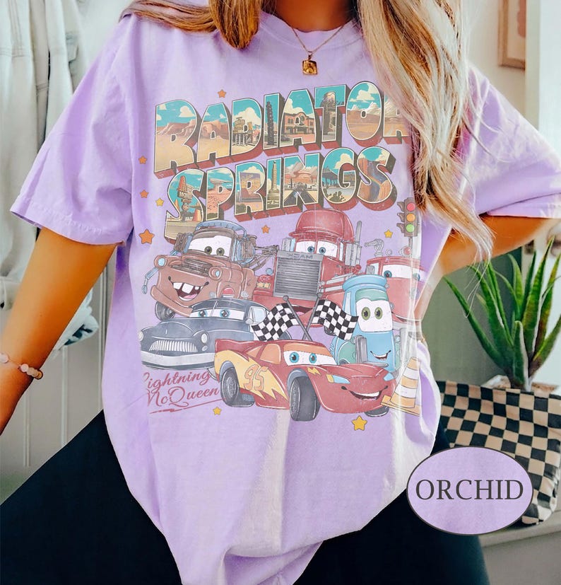 K&ouml;nnte beinhalten: Orchideenfarbenes T-Shirt mit einer Grafik von Charakteren aus dem Film Cars, darunter Lightning McQueen. Das Design enth&auml;lt den Text "Radiator Springs" und "Lightning McQueen". Das Shirt hat kurze &Auml;rmel und einen Rundhalsausschnitt.