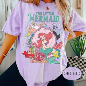 May include: Orchid-colored t-shirt featuring a graphic of Ariel from The Little Mermaid, surrounded by characters like Flounder, Sebastian, and Ursula. The shirt has the text "THE LITTLE MERMAID" in green. The shirt is a light purple color.
