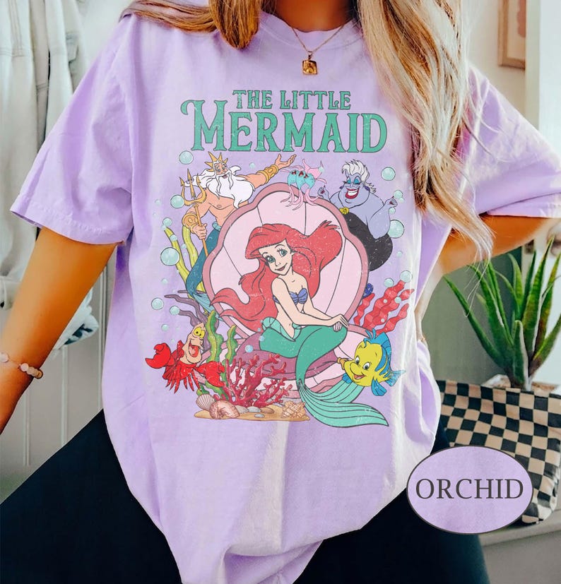 May include: Orchid-colored t-shirt featuring a graphic of Ariel from The Little Mermaid, surrounded by characters like Flounder, Sebastian, and Ursula. The shirt has the text "THE LITTLE MERMAID" in green. The shirt is a light purple color.